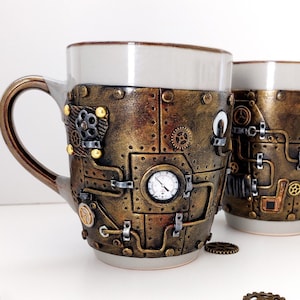 Pode incluir: Duas canecas de cerâmica com um design steampunk. As canecas são decoradas com tinta dourada, engrenagens e outros detalhes metálicos. Uma caneca tem um mostrador de relógio na frente.