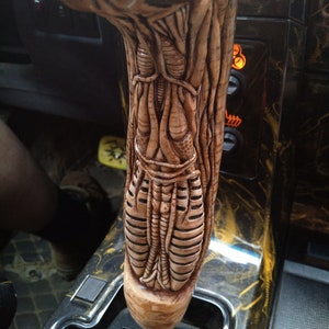 May include: A close-up of a custom car gear shift knob with a detailed, textured, alien-like design. The knob is made of a light brown material and is attached to a black gear shift lever.