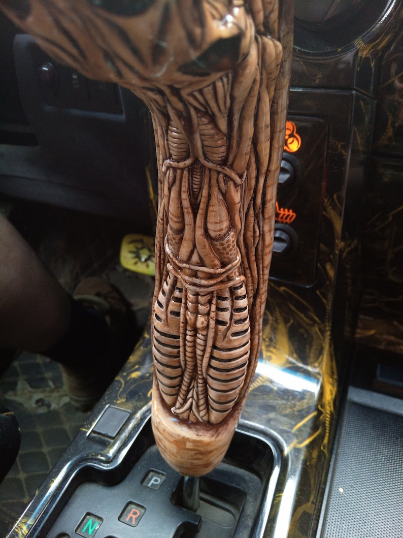 May include: A close-up of a custom car gear shift knob with a detailed, textured, alien-like design. The knob is made of a light brown material and is attached to a black gear shift lever.