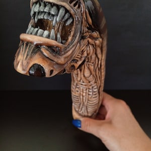 May include: A brown and black ceramic sculpture of an alien creature with a large open mouth and sharp teeth. The sculpture is detailed with a textured surface and appears to be a walking stick.