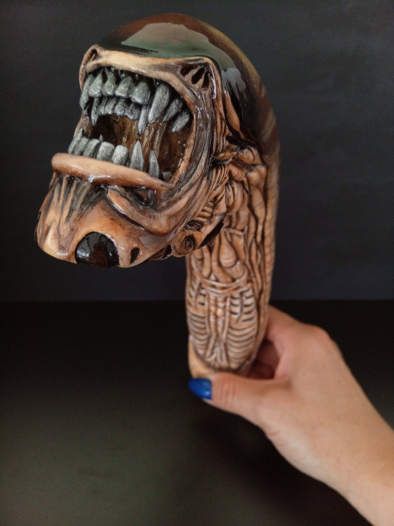 May include: A brown and black ceramic sculpture of an alien creature with a large open mouth and sharp teeth. The sculpture is detailed with a textured surface and appears to be a walking stick.