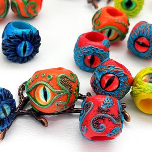 May include: A collection of colorful handmade polymer clay beads with eye designs. The beads are in various shades of red, orange, blue, green, and yellow. Some beads have intricate patterns and details.