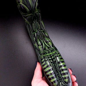 May include: A green and black alien-shaped sculpture with detailed anatomical features. The sculpture is made of a smooth, glossy material.