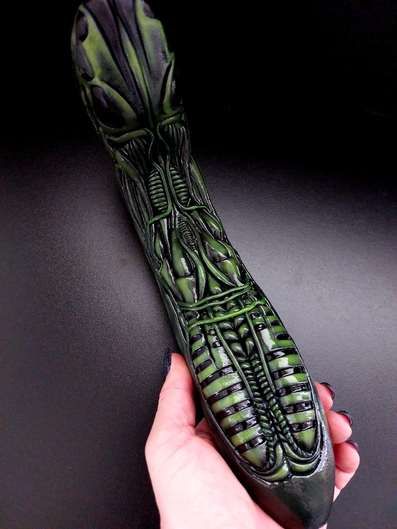 May include: A green and black alien-shaped sculpture with detailed anatomical features. The sculpture is made of a smooth, glossy material.
