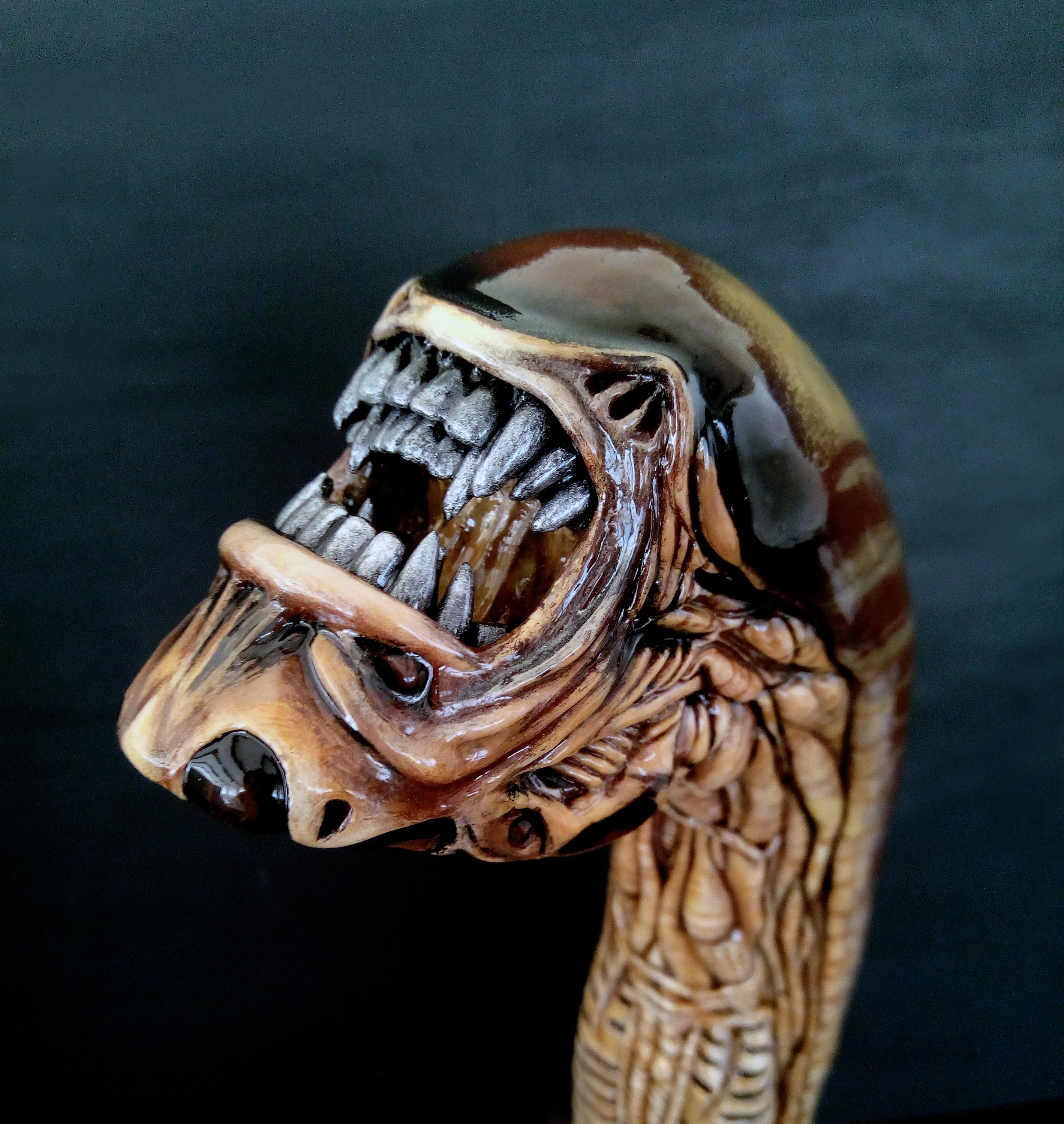 May include: A brown and beige wooden walking stick with a detailed carving of an alien creature's head with an open mouth and sharp teeth.
