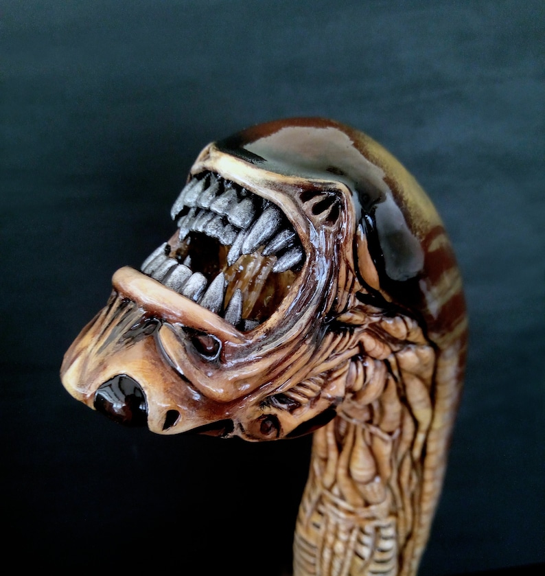 May include: A brown and beige wooden walking stick with a detailed carving of an alien creature's head with an open mouth and sharp teeth.