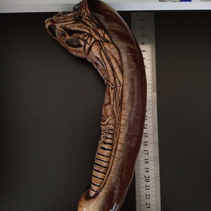 May include: A brown and beige alien creature sculpture, approximately 25 inches long, with detailed carvings. The sculpture is positioned against a black background with a ruler for scale.