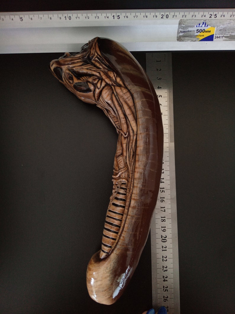 May include: A brown and beige alien creature sculpture, approximately 25 inches long, with detailed carvings. The sculpture is positioned against a black background with a ruler for scale.