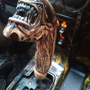 May include: A brown and black alien-shaped gear shift knob for a car. The knob is detailed with a textured, scaly design and a gaping mouth with sharp teeth.