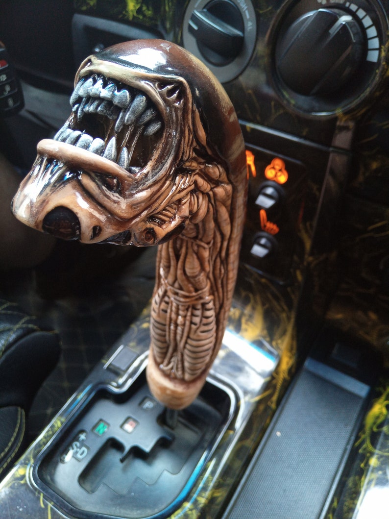 May include: A brown and black alien-shaped gear shift knob for a car. The knob is detailed with a textured, scaly design and a gaping mouth with sharp teeth.