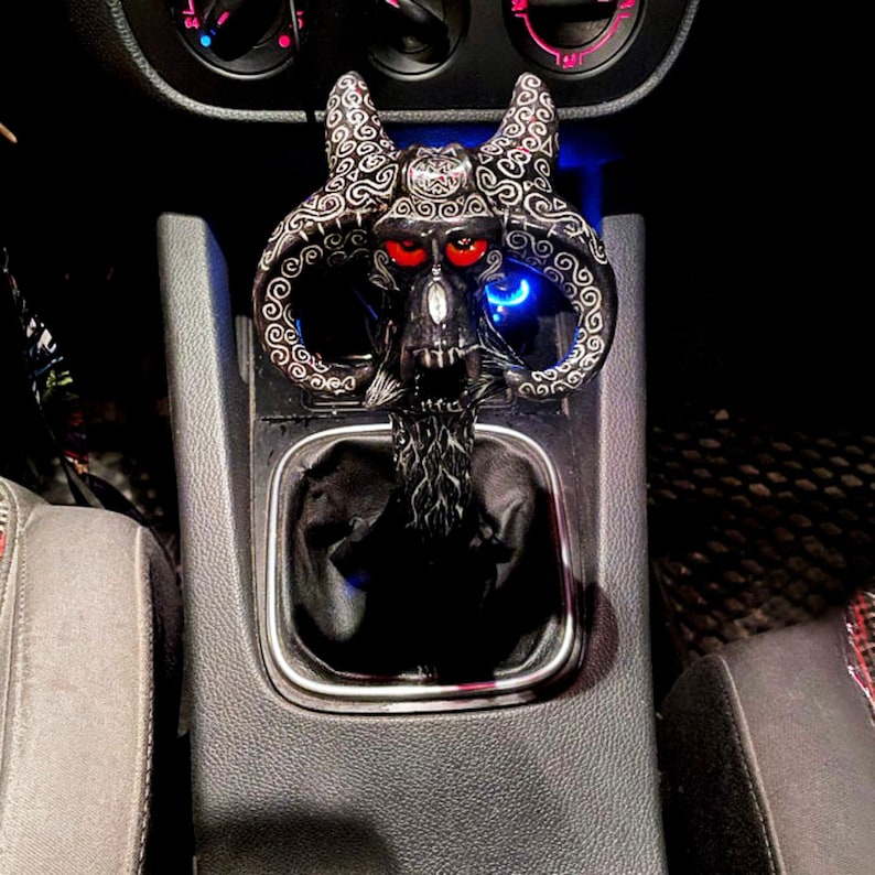Pu&ograve; includere: Un pomello del cambio a forma di teschio nero e argento con occhi rossi. Il pomello &egrave; installato nella console centrale di un'auto.