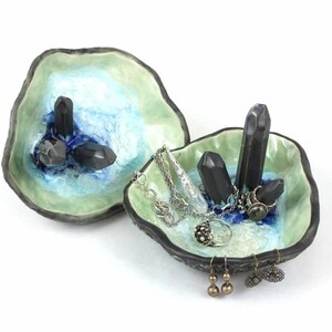 May include: A ceramic ring dish with a green and blue glaze. The dish is shaped like a seashell and has black crystal accents. The dish is filled with jewelry, including rings, earrings, and a necklace.