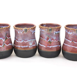 May include: Four handmade ceramic cups with a unique design. The cups feature a dark gray base, a burgundy and blue marbled pattern, and a decorative rim with small gold accents. Each cup has a small, dark gray stone detail.