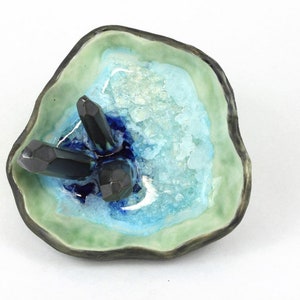 May include: A ceramic dish with a green, blue, and white glaze. The dish has a rough, organic shape and features three black crystal-like objects inside.