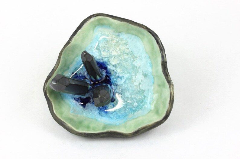 May include: A ceramic dish with a green, blue, and white glaze. The dish has a rough, organic shape and features three black crystal-like objects inside.