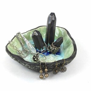 May include: A ceramic ring dish with a green and blue glaze. The dish features three black ceramic crystal-shaped holders. The dish is filled with jewelry, including rings, earrings, and necklaces.