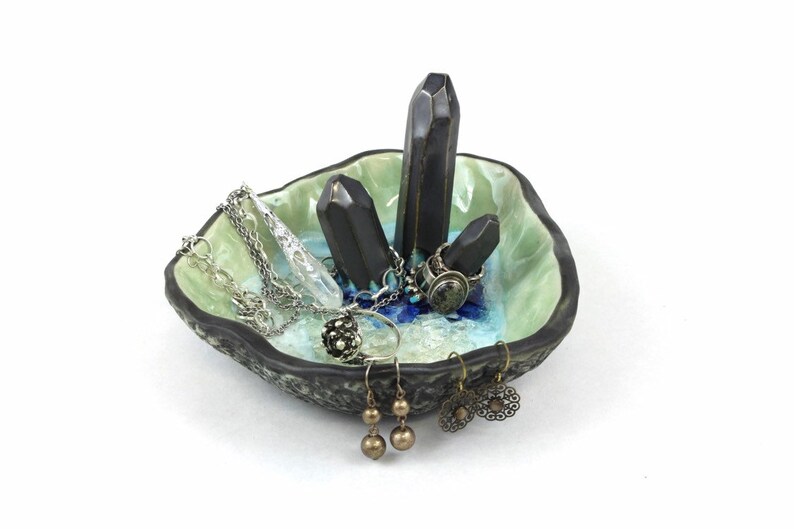 May include: A ceramic ring dish with a green and blue glaze. The dish features three black ceramic crystal-shaped holders. The dish is filled with jewelry, including rings, earrings, and necklaces.