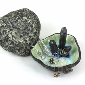 May include: A ceramic jewelry dish shaped like a seashell with a green and blue glaze. The dish has three black crystal-shaped holders and is filled with jewelry, including a silver chain necklace, a silver ring, and a pair of gold earrings.