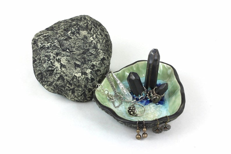 May include: A ceramic jewelry dish shaped like a seashell with a green and blue glaze. The dish has three black crystal-shaped holders and is filled with jewelry, including a silver chain necklace, a silver ring, and a pair of gold earrings.