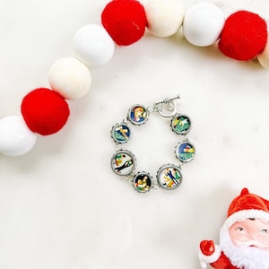 May include: A silver bracelet with a toggle clasp features eight round charms with colorful images of people and trees. The bracelet is displayed on a white surface with red and white pom poms.