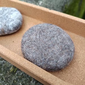 Może przedstawiać: Dwa szare, filcowane kamienie spoczywają w prostokątnej, jasnobrązowej tacy. Kamienie mają owalny kształt i teksturowany wygląd. Taca ma prosty, minimalistyczny design, a tło jest rozmyte, sugerując otoczenie na zewnątrz.
