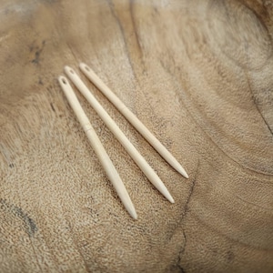 Op de afbeelding: Drie lichtbeige, gladde, langwerpige naainaalden met een puntig uiteinde en een klein, ovaalvormig oog aan de bovenkant. De naalden zijn in een parallelle, diagonale formatie gerangschikt en hebben een uniforme, slanke en taps toelopende vorm.