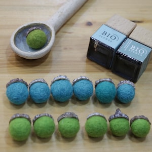 Peut inclure: Une cuillère en bois contient un gland en feutre vert. Deux petites bouteilles carrées avec des couvercles en bois sont étiquetées "BIO SCENTS". En dessous, des rangées de glands en feutre bleu et vert avec des chapeaux argentés, disposés sur une surface en bois.