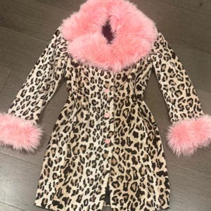 May include: A leopard print coat with a pink faux fur collar and cuffs. The coat has a button-down front and a classic silhouette. The leopard print features brown and black spots on a cream background, creating a bold and stylish look.