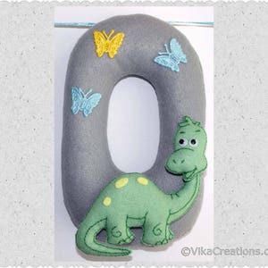 K&ouml;nnte beinhalten: Ein grauer Filzbuchstabe O mit einem gr&uuml;nen Dinosaurier und blauen und gelben Schmetterlingen. Der Dinosaurier hat ein l&auml;chelndes Gesicht und schaut hinter dem Buchstaben hervor.