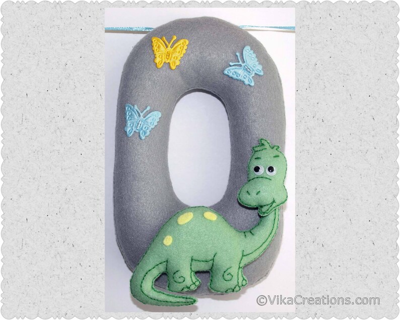 K&ouml;nnte beinhalten: Ein grauer Filzbuchstabe O mit einem gr&uuml;nen Dinosaurier und blauen und gelben Schmetterlingen. Der Dinosaurier hat ein l&auml;chelndes Gesicht und schaut hinter dem Buchstaben hervor.