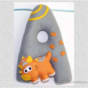 K&ouml;nnte beinhalten: Ein grauer Filzbuchstabe A mit einem gelben und orangefarbenen Dinosaurier-Design. Der Dinosaurier schaut hinter dem Buchstaben hervor. Der Buchstabe ist mit gelben Streifen und orangefarbenen Punkten verziert.