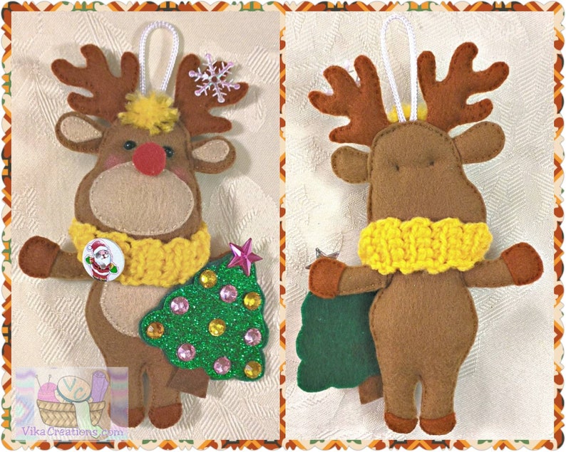 Puede incluir: Un adorno de reno de fieltro marr&oacute;n con una bufanda de punto amarilla, sosteniendo un &aacute;rbol de Navidad de fieltro verde con purpurina rosa y dorada. El reno tiene la nariz roja y un copo de nieve blanco en sus cuernos.