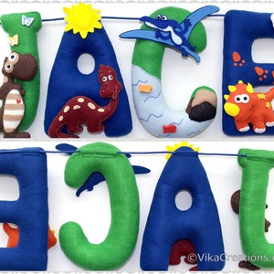 K&ouml;nnte beinhalten: Ein Filzbanner mit dem Namen "JACE" in blauen und gr&uuml;nen Buchstaben. Jeder Buchstabe ist mit einem anderen Dinosaurier-Charakter verziert, darunter ein Pterodactylus, ein Stegosaurus, ein Triceratops und ein Tyrannosaurus Rex. Das Banner ist mit einer blauen Schnur befestigt und hat einen wei&szlig;en Hintergrund.