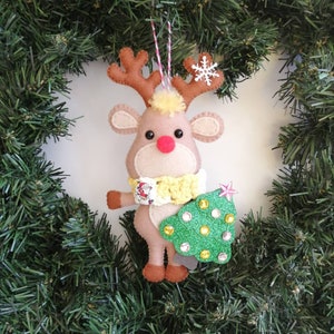 Puede incluir: Un adorno de reno de fieltro que sostiene un &aacute;rbol de Navidad de purpurina verde con una estrella rosa en la parte superior. El reno lleva una bufanda amarilla y blanca y tiene la nariz roja.