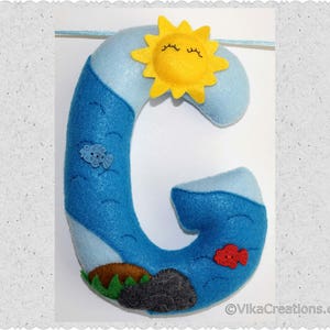 K&ouml;nnte beinhalten: Ein Filzbuchstabe G in Blau mit einer gelben Sonne und einem roten Fisch. Der Buchstabe ist mit wei&szlig;en Wolken und einer blauen Ozeanszene verziert.