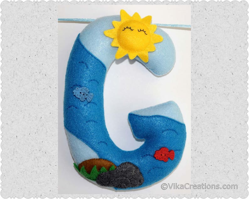 K&ouml;nnte beinhalten: Ein Filzbuchstabe G in Blau mit einer gelben Sonne und einem roten Fisch. Der Buchstabe ist mit wei&szlig;en Wolken und einer blauen Ozeanszene verziert.