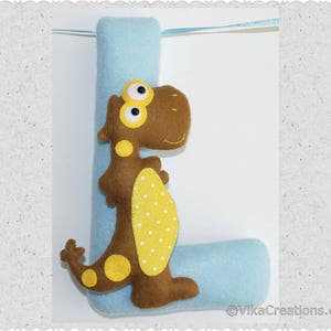 K&ouml;nnte beinhalten: Ein Filzbuchstabe L mit einem braunen Dinosaurier mit gelben Flecken daran. Der Buchstabe ist hellblau und der Dinosaurier hat gro&szlig;e Augen.