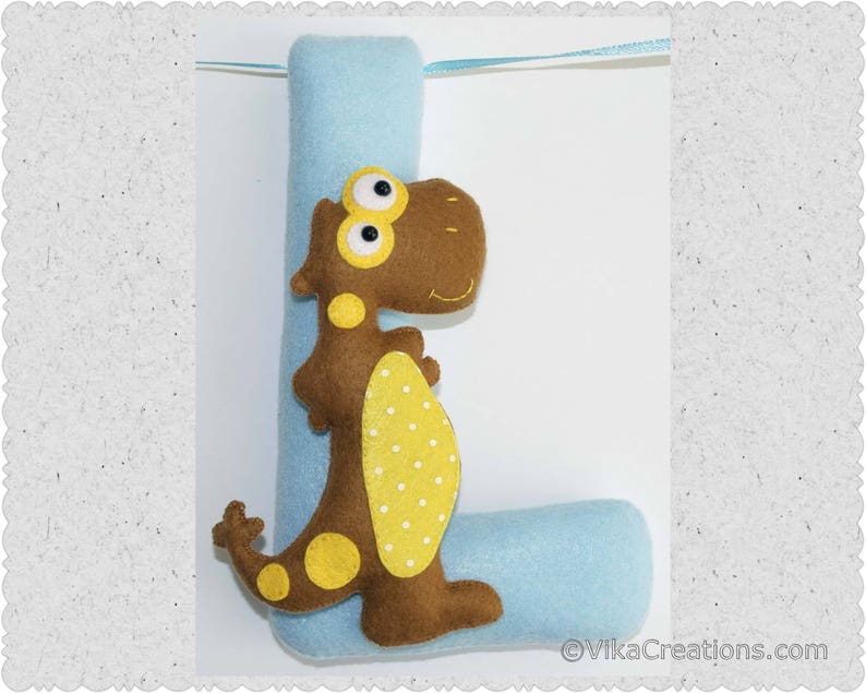 K&ouml;nnte beinhalten: Ein Filzbuchstabe L mit einem braunen Dinosaurier mit gelben Flecken daran. Der Buchstabe ist hellblau und der Dinosaurier hat gro&szlig;e Augen.