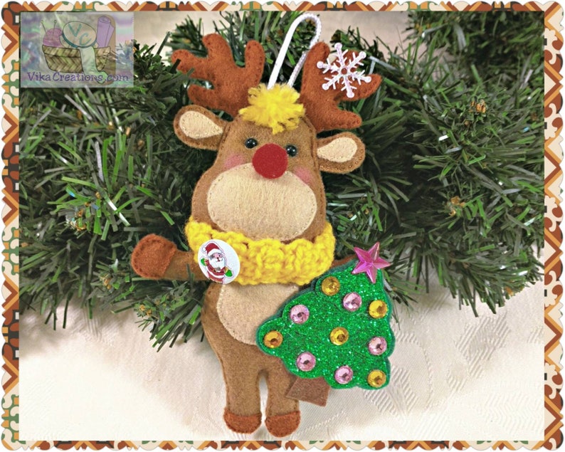 Puede incluir: Un adorno de reno de fieltro que sostiene un &aacute;rbol de Navidad de purpurina verde con adornos rosas y dorados. El reno lleva una bufanda amarilla y tiene la nariz roja.