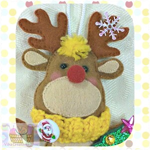 Puede incluir: Un adorno de reno de fieltro con una nariz roja, cuernos marrones y un pomp&oacute;n amarillo en la cabeza. El reno lleva una bufanda de crochet amarilla y un bot&oacute;n con una imagen de Pap&aacute; Noel.