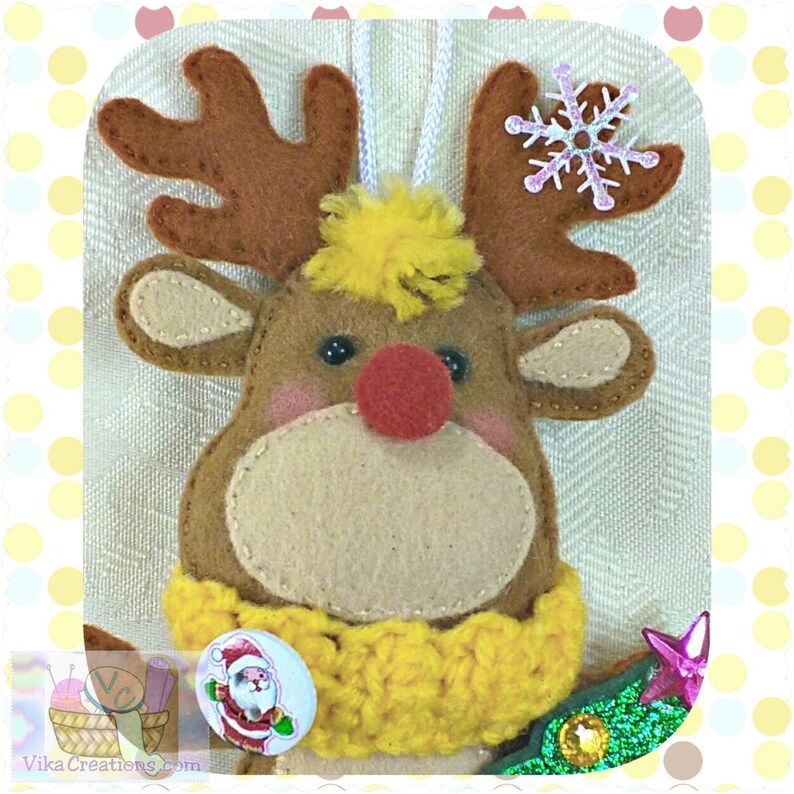 Puede incluir: Un adorno de reno de fieltro con una nariz roja, cuernos marrones y un pomp&oacute;n amarillo en la cabeza. El reno lleva una bufanda de crochet amarilla y un bot&oacute;n con una imagen de Pap&aacute; Noel.