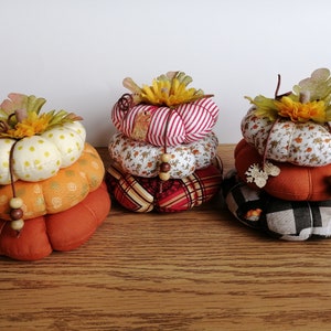 May include: Three stacked fabric pumpkins with different patterns. The top pumpkin is white with yellow polka dots, the middle pumpkin is orange with a floral pattern, and the bottom pumpkin is orange with a brown and white pattern. Each pumpkin has a brown stem and a green leaf.