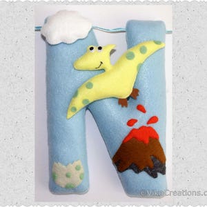 K&ouml;nnte beinhalten: Blauer Filzbuchstabe N mit einem gelben Dinosaurier, einem roten Vulkan und einer wei&szlig;en Wolke. Der Buchstabe ist mit gr&uuml;nen und wei&szlig;en Punkten verziert.