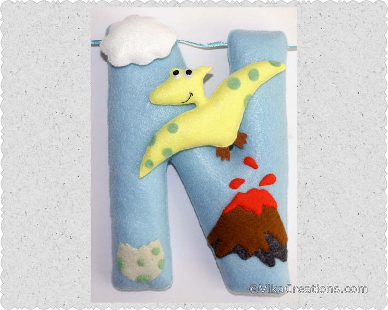 K&ouml;nnte beinhalten: Blauer Filzbuchstabe N mit einem gelben Dinosaurier, einem roten Vulkan und einer wei&szlig;en Wolke. Der Buchstabe ist mit gr&uuml;nen und wei&szlig;en Punkten verziert.