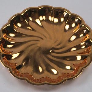 May include: A shiny, gold-colored decorative plate with a scalloped edge. The plate has a spiral design in the center and a decorative border. The plate is made of a reflective material.