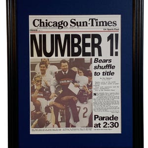 May include: A framed Chicago Sun-Times newspaper front page from January 27, 1986, featuring the headline NUMBER 1! and Bears shuffle to title. The black and white photograph shows football players celebrating, with a dark blue mat and black frame.