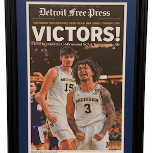 May include: Framed Detroit Free Press newspaper front page celebrating the Michigan Wolverines' 2026 NCAA National Championship victory. The headline reads "VICTORS!" with images of celebrating basketball players in maize and blue uniforms.