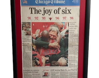 May include: A framed Chicago Tribune newspaper front page from June 15, 1998, titled The joy of six. The print features a photograph of Michael Jordan in a red jersey embracing coach Phil Jackson, with a red matting and a black frame.