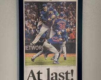 Pode incluir: Página de jornal emoldurada do Chicago Tribune celebrando a vitória do Chicago Cubs na World Series. A manchete "At last!" é proeminente, com uma foto de jogadores celebrando. A moldura preta realça a exibição.
