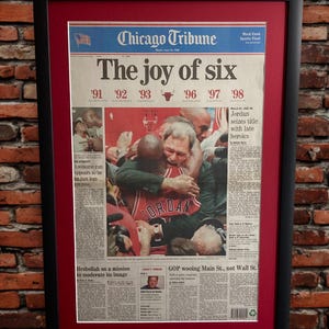 May include: Framed Chicago Tribune newspaper front page titled "The joy of six" with a photo of a basketball player. The frame is black with a red matting. The newspaper is from June 13, 1996.